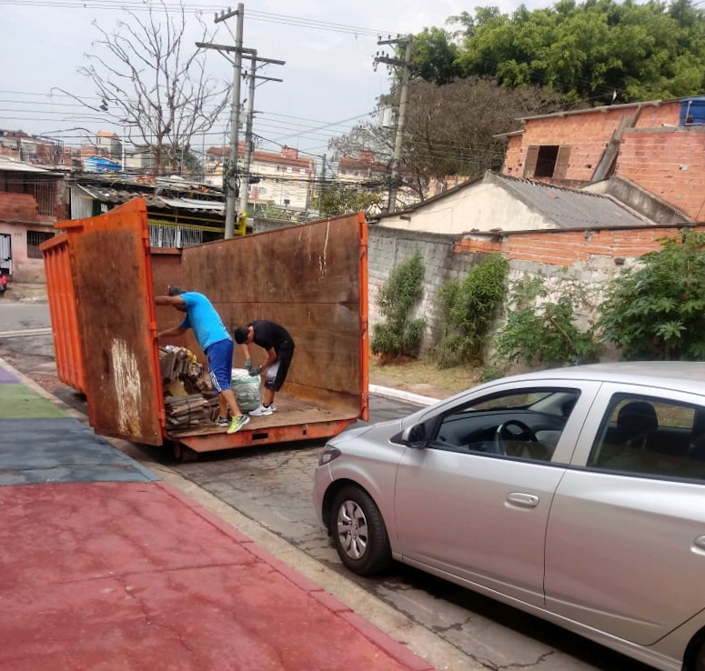 Em uma rua de bairro com casas simples e vegetação ao fundo, dois homens estão dentro de uma grande caçamba metálica laranja, organizando materiais descartados, como papelão e sacos com resíduos. Um deles usa roupa esportiva azul, enquanto o outro veste preto. Um carro prata está estacionado próximo à caçamba, e o chão ao redor tem áreas coloridas, possivelmente de um espaço público ou playground. A cena ilustra um descarte adequado de resíduos em local apropriado.